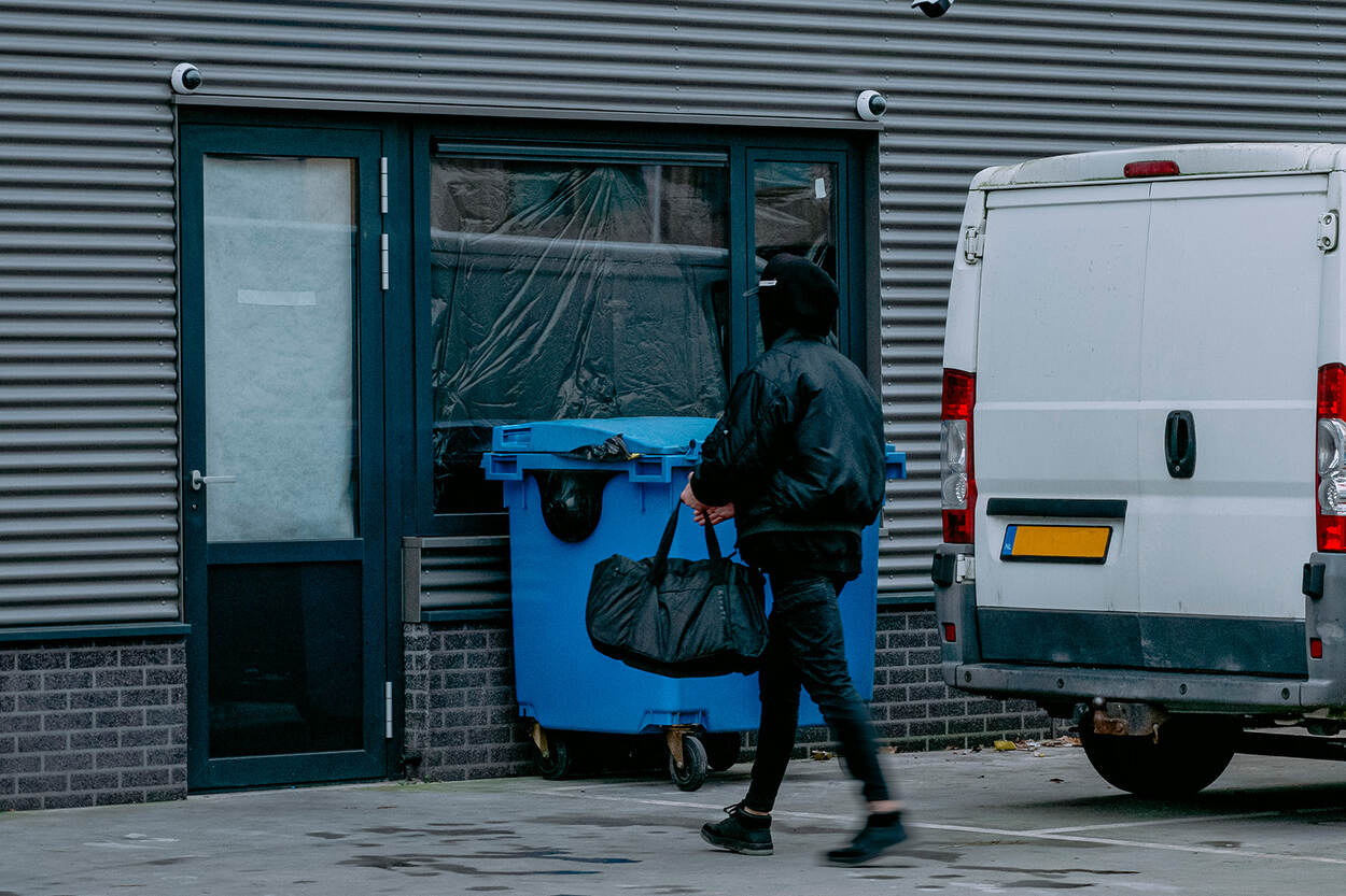 Iemand loopt vanaf een anoniem, wit busje naar een bedrijfsgebouw. Hij draagt een capuchon over zijn hoofd en heeft een grote sporttas vast. Het gebouw heeft afgeplakte ramen.