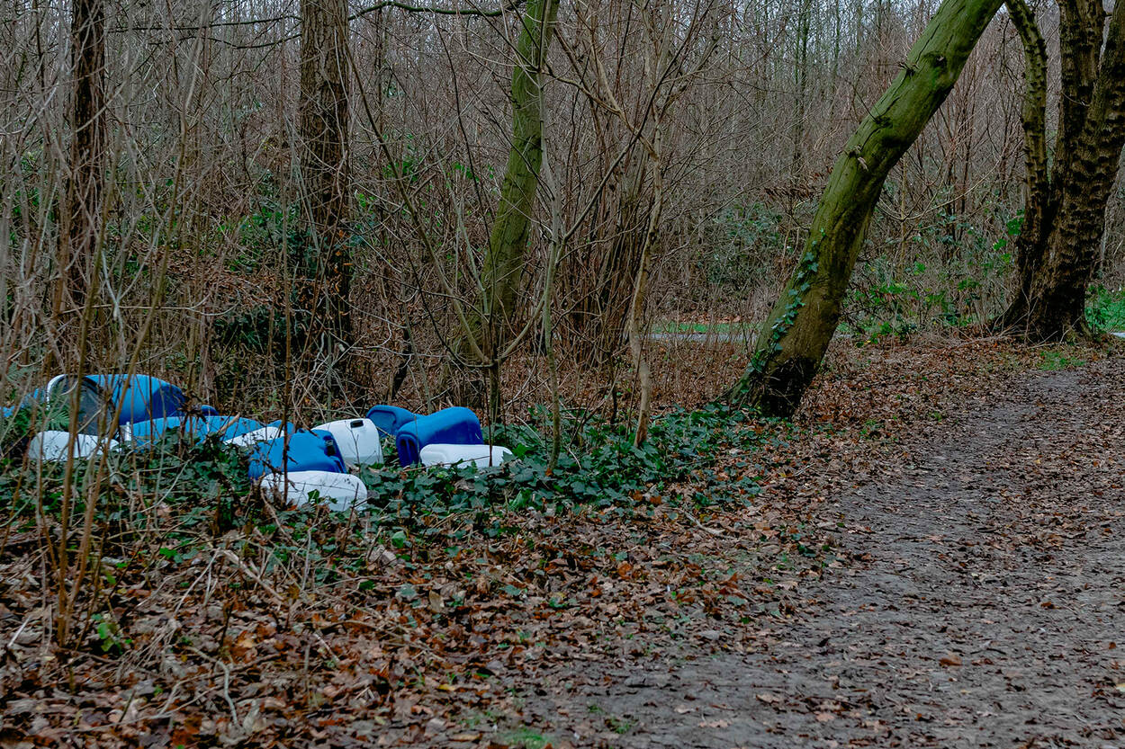 Foto van een plek in een bos, waar tussen de bomen en struiken lege blauwe en witte jerrycans liggen.