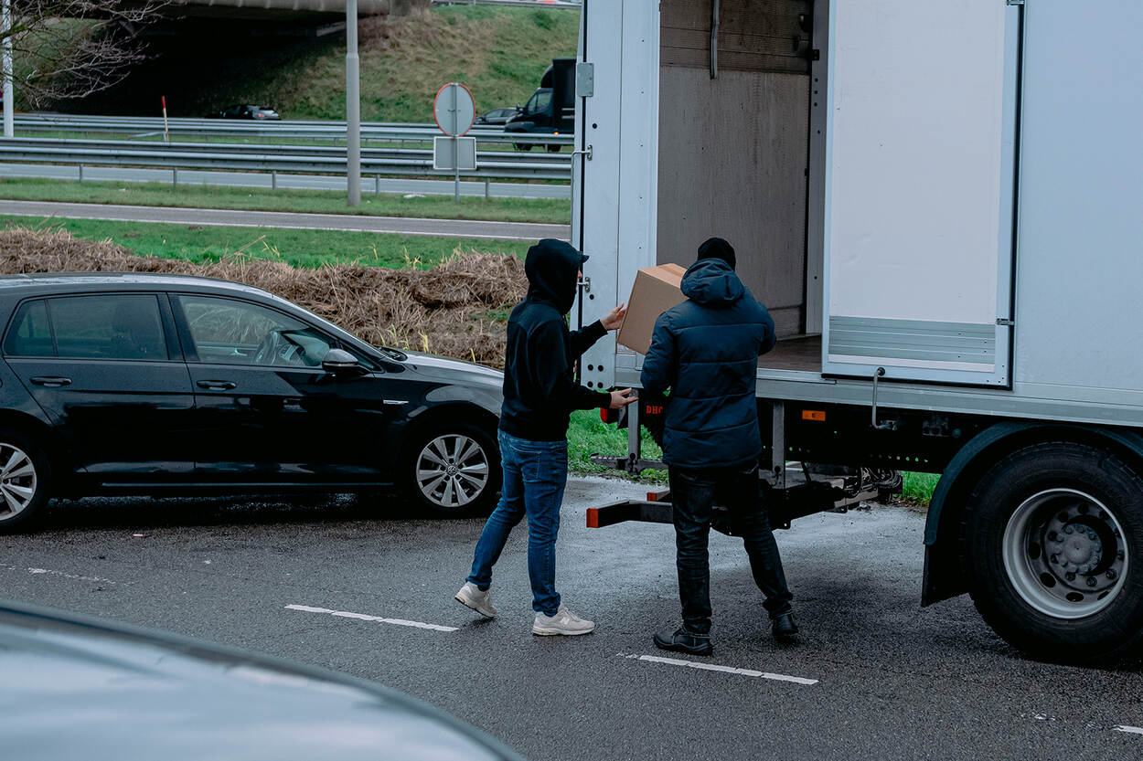 Een vrachtwagen staat stil op een normale autoparkeerplaats. Iemand met een capuchon geeft een pakketje aan een ander, om in de laadruimte van de vrachtwagen te stoppen.